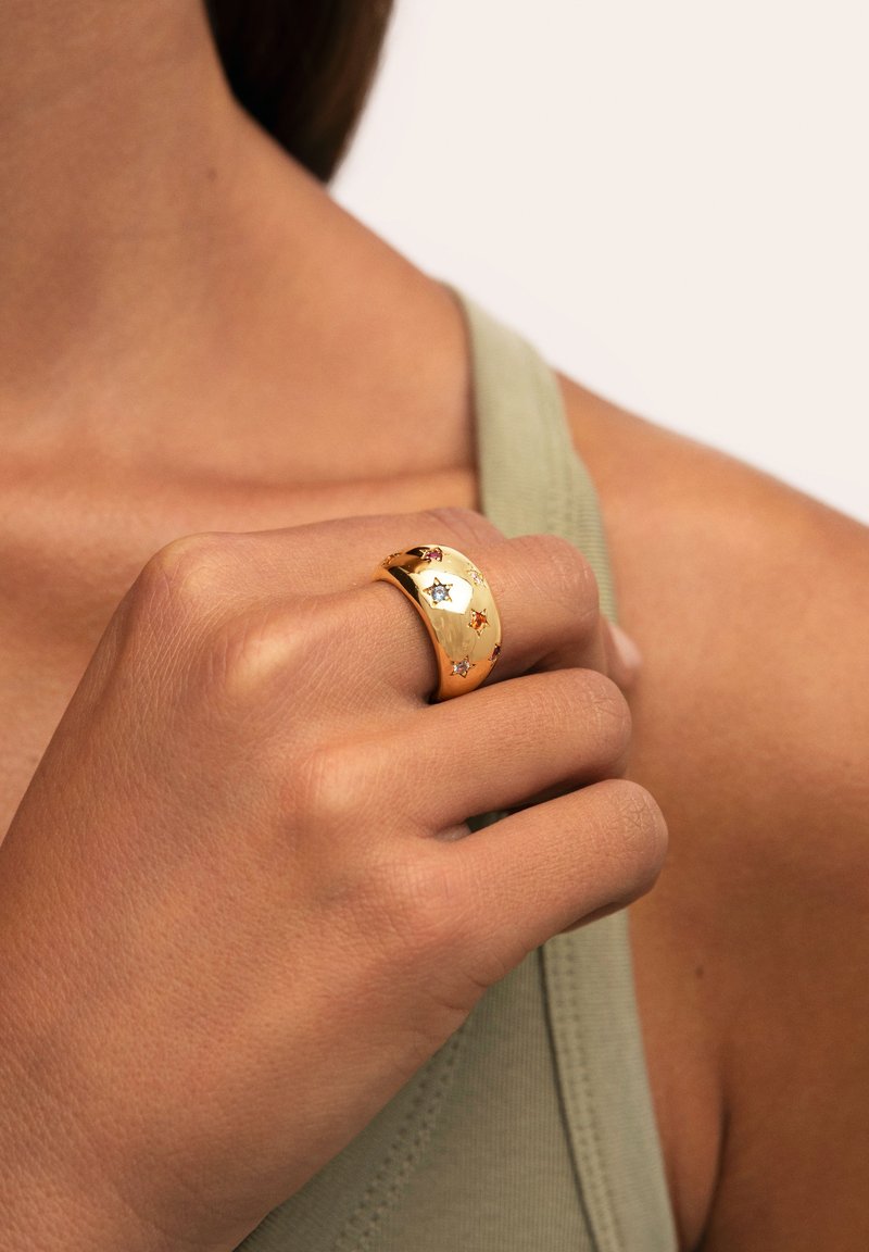 Anillo de banda de oro con motivos de estrellas en acentos rojos, azules y transparentes. Superficie lisa y brillante, llevado en una mano contra una blusa verde apagado.
