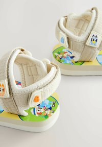 Sandalias para niños con correas de tela beige, suelas blancas y gráficos de dibujos animados coloridos en la plantilla. Cierre de velcro para facilitar el uso.