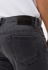 Gray denim jeans featuring a black leather waistband label with "Hi-Flex" and "Brax" branding. Includes back pockets with subtle stitching.