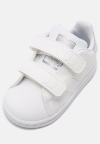 White synthetic sneaker with two Velcro straps, rounded toe, textured surface, logo on tongue, and perforated details on the sides.