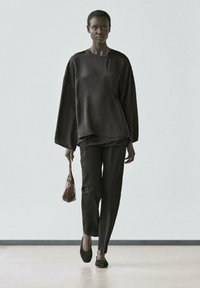 Top nero oversize con scollo a barca, abbinato a pantaloni neri slim. La modella tiene in mano una borsa marrone e indossa ballerine nere.