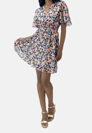 Kvinde iført en marineblå blomsterkjole med orange og hvide blomster, flagrende ærmer og guld høje hæle sandaler, der holder kjolens kant.