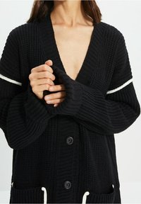 Svart stickad cardigan med djup V-ringning, ribbad textur och långa ärmar. Har vita accentränder och knappstängning.