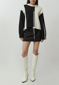 Pull cropped en tricot noir et crème avec un panneau texturé et des détails sur les côtés, associé à une mini-jupe noire et des bottes blanches jusqu'aux genoux.