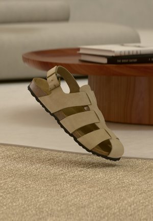 Bēšs zamšādas sandales ar trim siksnām, sprādzi, korķa zolīti, melnu protektoru, peldoši virs austas bēšas paklāja, koka kafijas galdiņa un dīvāna fonā.