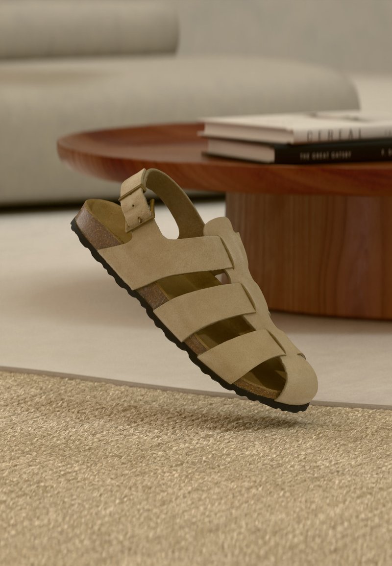 Sandale en daim beige avec trois bandes, boucle, semelle en liège, semelle extérieure noire, flottant au-dessus d'un tapis beige tissé, table basse en bois et canapé en arrière-plan.