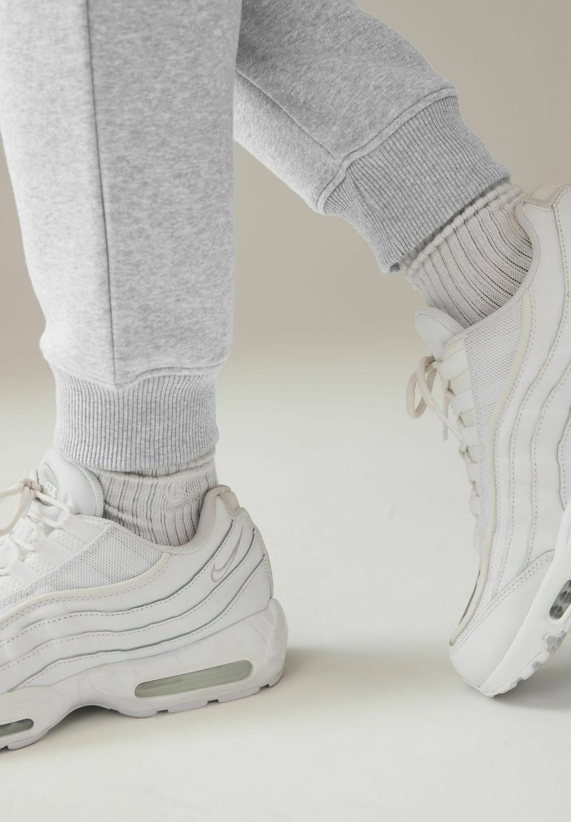 Weiße Sportsneaker mit Mesh- und Lederdetails, getragen mit hellgrauen gerippten Socken. Graue Jogginghosen mit elastischen Bündchen sind sichtbar.