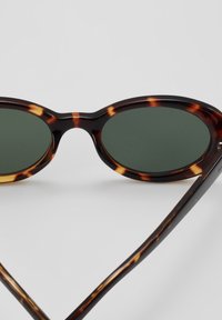 Gafas de sol de carey con lentes ovalados verdes. Montura gruesa y brillante con un patrón de marrón oscuro y claro. Patillas finas y curvadas.