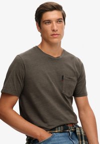 Szara koszulka z krótkim rękawem wykonana z miękkiej bawełny, z kieszonką na piersi z metką, okrągłym dekoltem i luźnym krojem.