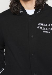 Černá bunda ve stylu varsity z bavlněné směsi, s bílým vyšitým logem, cvočky a kontrastním límcem a manžetami s proužky.