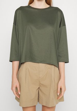 Femme portant un haut ample à manches trois-quarts de couleur vert olive et un short plissé taille haute beige, debout devant un fond uni.