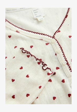 Långärmad, lätt krämfärgad topp med maroonhjärtmönster och texturerade ränder. Den har knappstängning och volanger längs halsringningen.