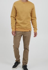 Bluza w kolorze musztardowym z okrągłym dekoltem, ściągaczami przy rękawach i u dołu. W połączeniu z jasnobrązowymi spodniami chinos i czarnymi tenisówkami. Gładka faktura.