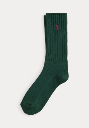 Chaussette côtelée vert foncé avec un petit logo rouge brodé près du bord sur un fond clair uni.