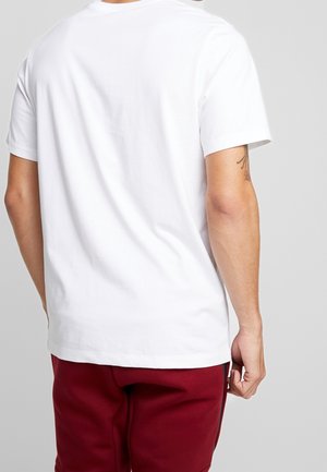 Camiseta blanca de algodón con cuello redondo y mangas cortas, vista desde la parte posterior. Combinada con pantalones de chándal rojos. Diseño simple, sin patrones visibles.