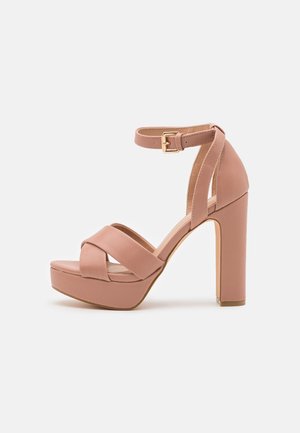 Blush-pink High-Heel-Sandale mit Plateausohle, überkreuzten Vorderriemen, Knöchelriemen und goldener Schnalle auf einem weißen Hintergrund.