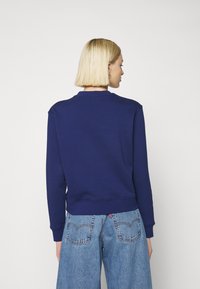 Tamsiai mėlynas megztinis su apvalia iškirpte, pyniais rankogaliais ir apačia. Dėvimi su šviesiai mėlynomis plačiomis kelnėmis, turinčiomis klasikines galines kišenes.