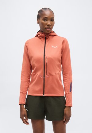 Kvinde iført en koralfarvet hættetrøje med lynlås og brystlomme samt sorte shorts, stående mod en ensfarvet lys baggrund.