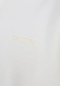 Weißer Stoff mit hellbeigem, gesticktem Text "SMILODOX". Die glatte Textur hat einen leichten Glanz, die Nahaufnahme hebt die Materialqualität hervor.