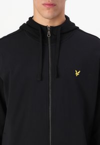Sudadera negra con cremallera y un solo logo de un pájaro amarillo en el pecho, con capucha ajustable y textura de tela suave.
