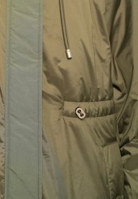 Groene jas met een cinchkoord en metalen toggles op de taille, getoond in close-updetail van de stof en de stiksels.