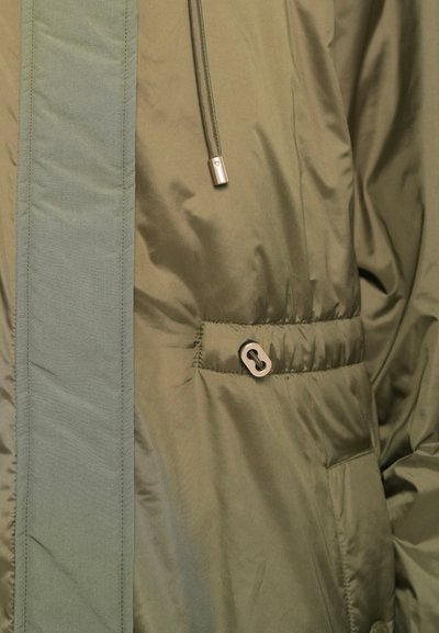Green jacket with a cinch cord and metal toggle on the waist, shown in close-up detail of fabric and stitching.