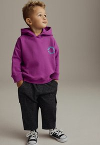 Lila Sweatshirt mit vorderem Kängurutasche, versehen mit dem Text "Big Smiles". Schwarze gemusterte Hose, weiße Turnschuhe mit schwarzen Akzenten.