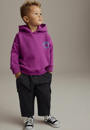 Lila Sweatshirt mit vorderem Kängurutasche, versehen mit dem Text "Big Smiles". Schwarze gemusterte Hose, weiße Turnschuhe mit schwarzen Akzenten.
