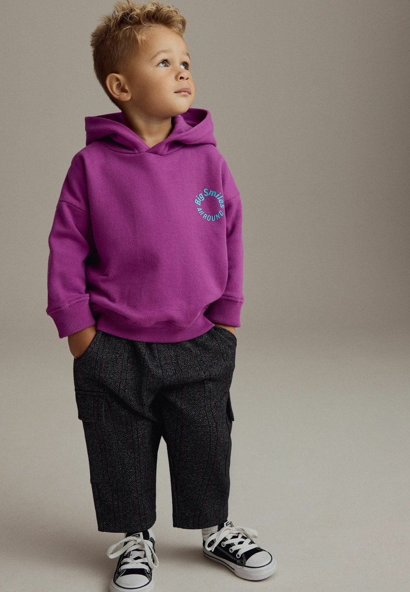 Lila Sweatshirt mit vorderem Kängurutasche, versehen mit dem Text "Big Smiles". Schwarze gemusterte Hose, weiße Turnschuhe mit schwarzen Akzenten.