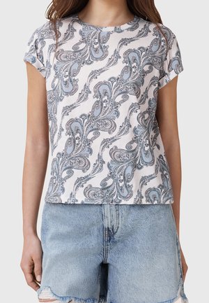 Frau trägt ein weißes kurzärmeliges Hemd mit blauem Paisley-Muster, kombiniert mit hellblauen, ausgewaschenen Jeansshorts.