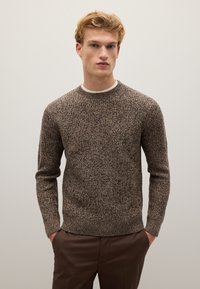Maglione a maglia marrone con texture, collo a giro e maniche lunghe, caratterizzato da un orlo a coste, indossato su una camicia bianca.
