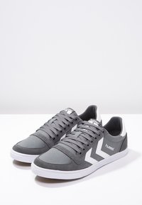 Sneakers in tela grigia con accenti bianchi, caratterizzati da una punta arrotondata, una chiusura con lacci e una superficie testurizzata. Logo del marchio visibile sul lato.