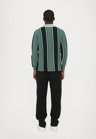Grüner und schwarzer gestreifter Pullover mit langen Ärmeln und einem weißen Kragen, kombiniert mit schwarzen Hosen und weißen Sneakern.