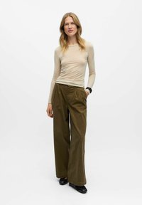 Beige langærmet bluse med en tætsiddende pasform og subtil tekstur; højtaljede olivengrønne vide bukser med folder; sorte sko.