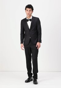 Čierny smoking s saténovým reverzom, biela košeľa a čierna motýlik. Čierne topánky dopĺňajú formálny outfit. Jednoduchý, upravený strih.