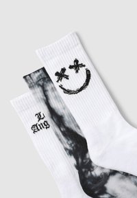 Trzy pary skarpetek: dwie białe z czarną uśmiechniętą buźką i napisem oraz jedna w czarno-białym wzorze tie-dye. Na tkaninie widoczne prążki.