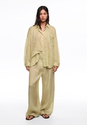 Mujer con camisa holgada de botones texturizada de color amarillo claro y pantalones anchos a juego, con sandalias negras, de pie sobre un fondo blanco.