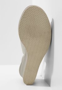Semelle d'une basket beige avec un motif ondulé, positionnée à la verticale contre un mur d'angle blanc sur une surface blanche.