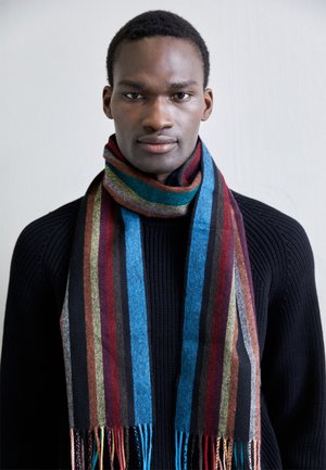 Écharpe en laine avec des rayures verticales noires, bleues, rouges et vertes, bordée de franges. Portée de manière décontractée sur un pull côtelé noir.