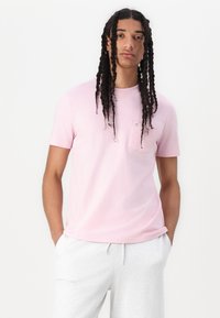 T-shirt rose clair à manches courtes avec une poche à l'avant, en coton, coupe décontractée, associé à un pantalon de survêtement gris. Design simple et minimal.