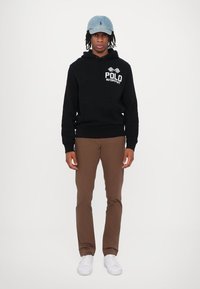 Melna kapuce džemperis ar "POLO MOTORSPORT" logo, brūnas bikses un gaiši zila denima cepure. Balti snekeri noslēdz apģērbu.