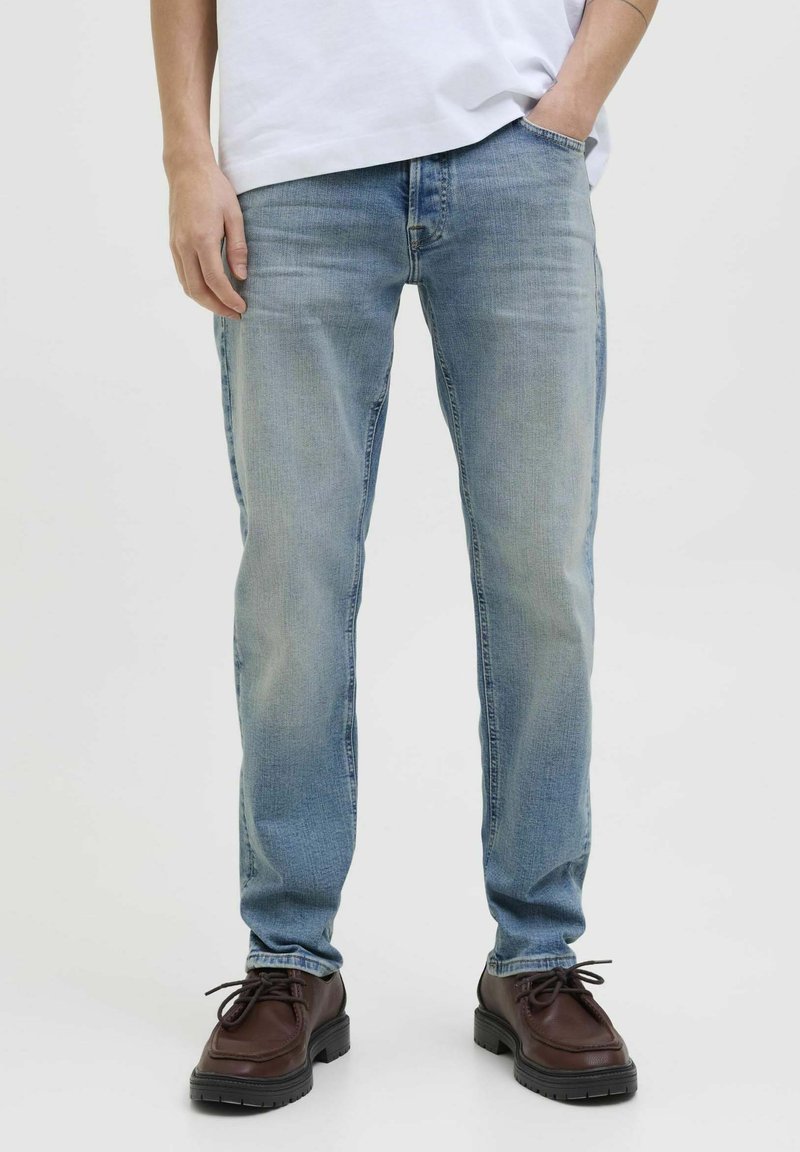 Jean en denim bleu clair avec une coupe droite, présentant un léger délavage et une texture lisse. Associé à des chaussures en cuir brun foncé à lacets.