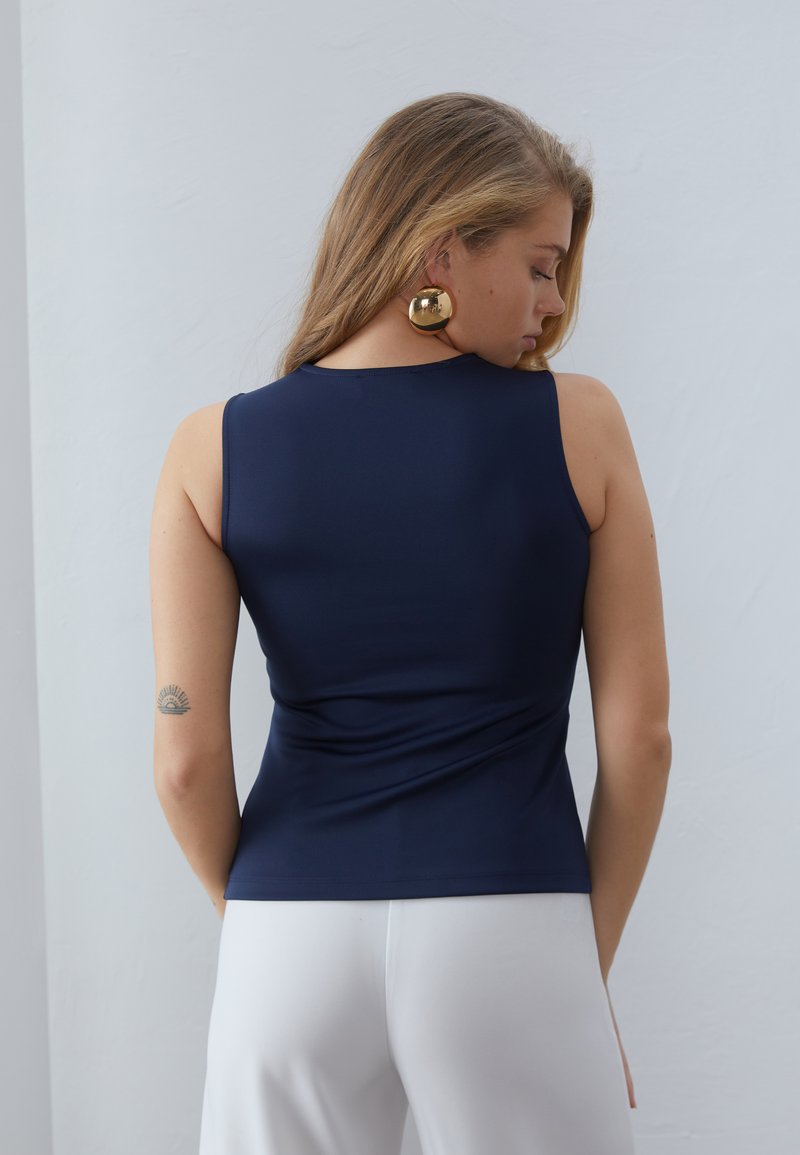 Granatowy bezrękawnik o gładkiej fakturze i dopasowanym kroju, z plecami w splot. W zestawieniu z białymi spodniami.