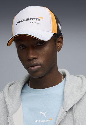 Jonge man met een McLaren Racing pet en een lichtblauw Puma McLaren shirt, met een lichtgrijze zip-up hoodie tegen een grijze achtergrond.