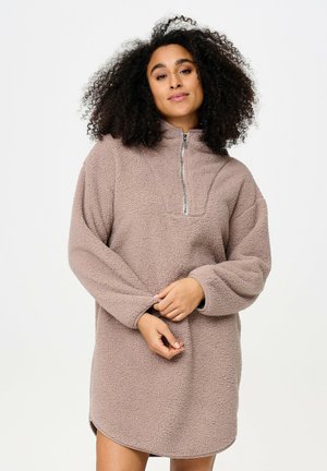 Femme aux cheveux bouclés portant une robe-pull polaire longue, beige et texturée, avec une fermeture éclair devant, debout devant un fond blanc uni.