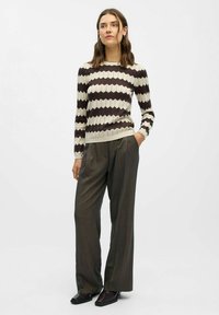 Strickpullover mit einem cremefarbenen und dunkelbraunen Zickzackmuster, kombiniert mit hoch geschnittenen grauen Hosen und schwarzen strukturierten Schuhen.