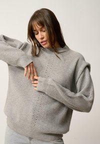 Maglione lavorato a maglia grigio chiaro con texture a coste, caratterizzato da un collo alto, spalle scese e dettagli verticali sottili lungo la parte anteriore.