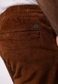 Pantalones de pana marrón con una textura acanalada, que cuentan con un bolsillo trasero y una pequeña etiqueta de metal de la marca. Tiro relajado y cintura elástica.