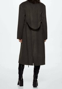 Manteau en laine vert foncé à coupe droite, manches longues, avec un détail ceinture à l'arrière, associé à des bottes noires montantes jusqu'aux genoux.