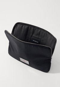 Funda de nailon negra con cremallera, de textura suave, que presenta una placa de logo plateada y una pequeña etiqueta negra en su interior. Diseño minimalista.